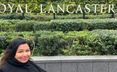 Tanvi is a brown powered-wheelchair user with long brown hair. She sits smiling in front of a large green bush with the words Royal Lancaster embedded in white cross the front of it