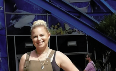 Natalie, a white female with blonde hair is pictured in front of Sea Life Birmingham. She is smiling at the camera and is wearing a black vest top and khaki green dungaree dress.