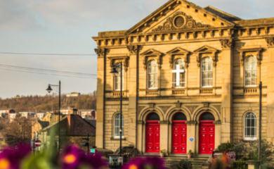 The front of Batley Methodist Church. A large stone building with 3 big red doors and many windows around the building. Close up flowers in the front of the image blurred out and the grey sky above.