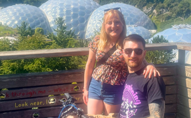 Becky and her partner Dan. Becky is standing and Dan is seated on his electric wheelchair. They are smiling and the domes of the Eden Project are in the background.