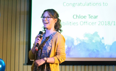 Chloe, a young white woman with brown hair and dark glasses, is giving a speech on stage. Behind her it says Chloe Tear Disabilities Officer 2018/19