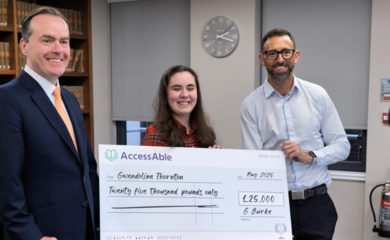 Gregory Burke (left) a white male in a dark blue suit poses for a photo with Gwendoline Thornton (middle) holding a large cheque that reads ‘Gwendoline Thornton’ A camera out of focus in the foreground. 