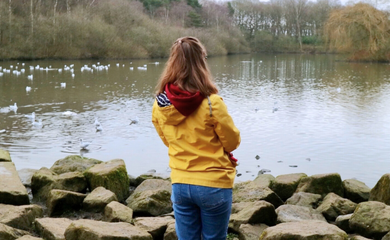 Chloe, a white woman with brown hair has her back to the camera. She’s wearing a yellow coat, blue jeans and is holding a long white cane. She’s stood by a lake 