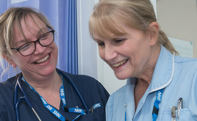 Image shows two women in dark blue and light blue NHS uniform smiling together. 