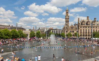Image of Bradford centre, there is a large water fountains in the middle and people around the water. There are buildings in the background. 