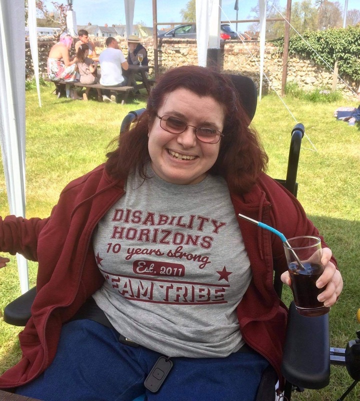 Emma Purcell as an adult, a white female who is a wheelchair user. She is smiling, wearing glasses and a grey Disability Horizons t-shirt with a red hoodie.