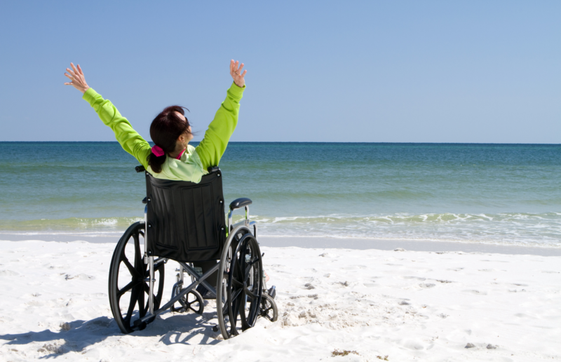 A white female is sat in her manual wheelchair on the beach. The picture is taken from behind her. Her black hair is in a ponytail and she is wearing a lime green long sleeved top. Her hands are stretched up into the air. The sea is visible in the near distance. The wheelchair has cast a shadow on the sand.