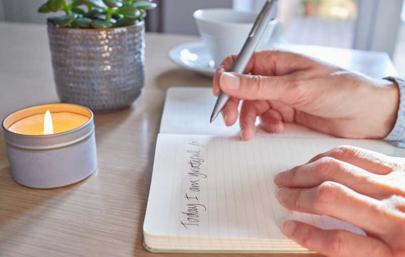 The image shows the top of a light coloured wooden table. On the far left of the image, there is a small, lit candle. There is also a plant in a grey, textured pot and a white teacup and saucer on the table. In the foreground, a person holding a silver pen is writing in a lined notebook. They have written 'Things I Am Grateful For' at the top of the page.