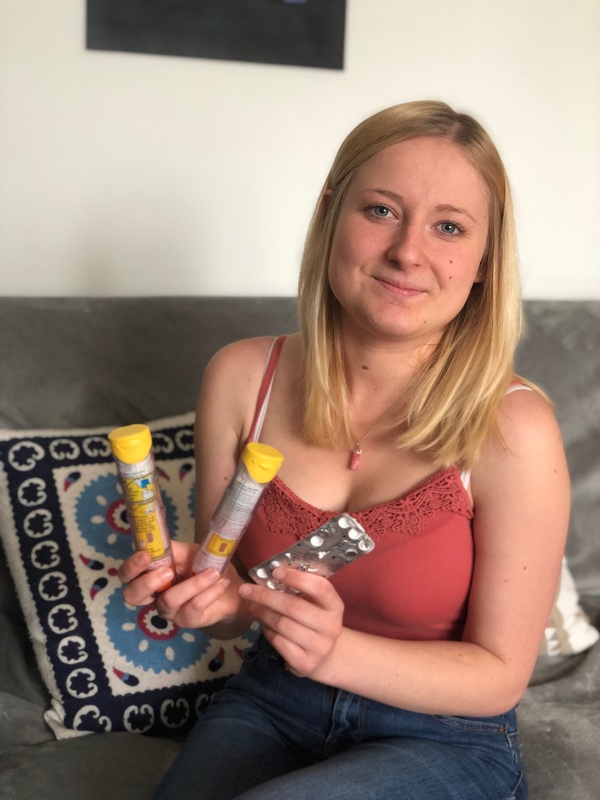 Becky is a white woman with long blonde hair. She is pictured sat down in a rust coloured vest top. She is holding two EpiPens and a strip of antihistamine tablets.