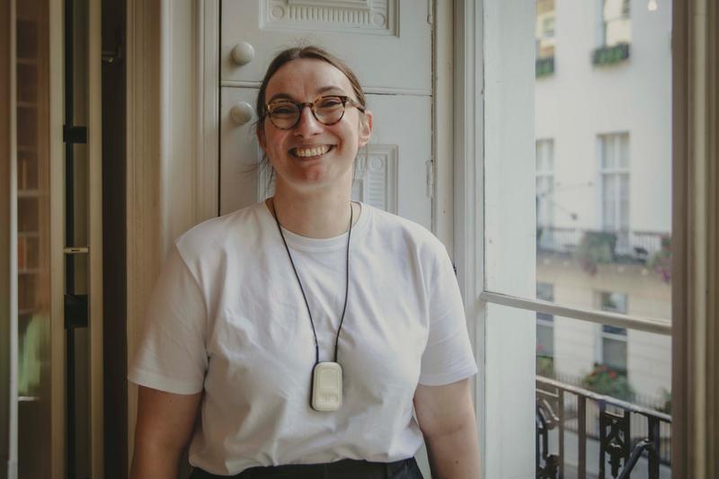Natasha is a white woman with brown hair, she is standing next to an open window smiling. She is wearing a white t-shirt, glasses, hearing aids and an assistive listening device around her neck.