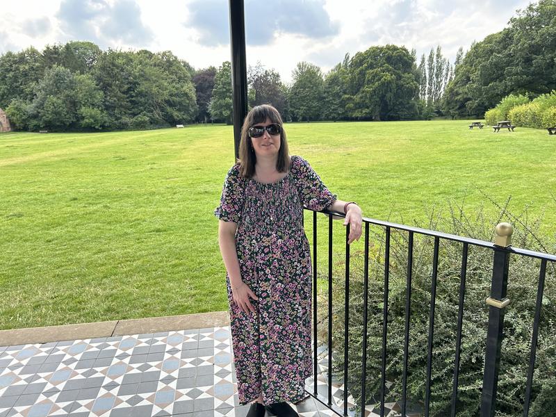 Holly stands on a tiled terrace in a large green park, leaning one arm on a black metal railing. She wears dark sunglasses and a long floral dress with short sleeves. A wide lawn stretches out behind her with trees in the distance and picnic benches scattered across the grass. The sky is partly cloudy and the scene feels bright and open.