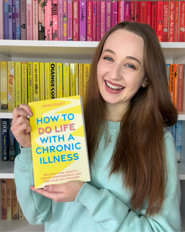 Pippa smiles at the camera while holding up a yellow book titled How to Do Life with a Chronic Illness by Pippa Stacey. She has long brown hair and wears a light mint green top. Behind her is a bookshelf arranged in colour order, with rows of bright pink, yellow, and orange book spines creating a vivid background.