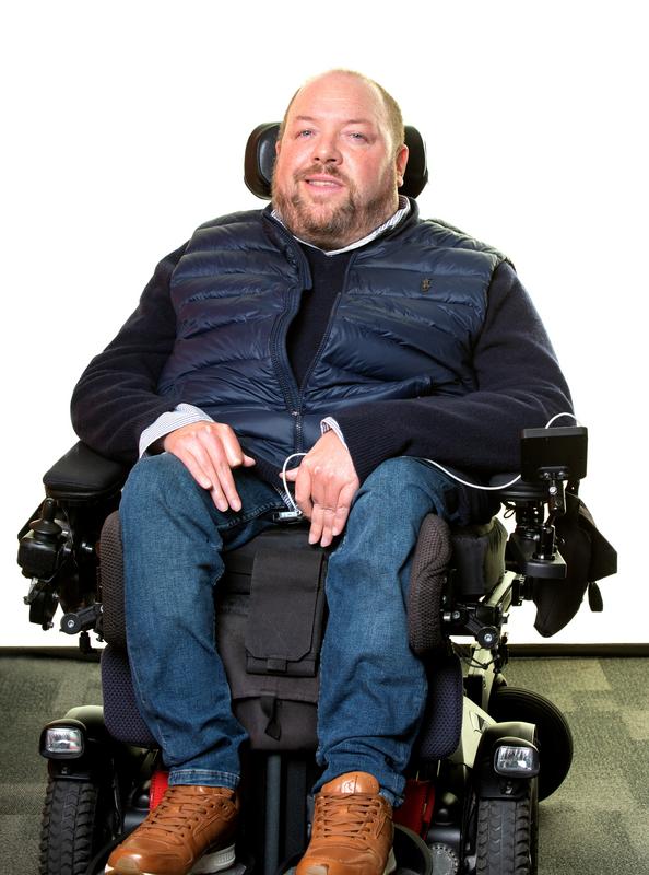 Ross sits facing the camera in a powered wheelchair against a plain white background. He has short hair and a trimmed beard and wears a navy jumper, a padded dark blue gilet, blue jeans, and brown trainers. His hands rest on his lap, and the wheelchair’s controls and supports are visible at the sides. He is smiling gently, and the image has a clean studio style feel.