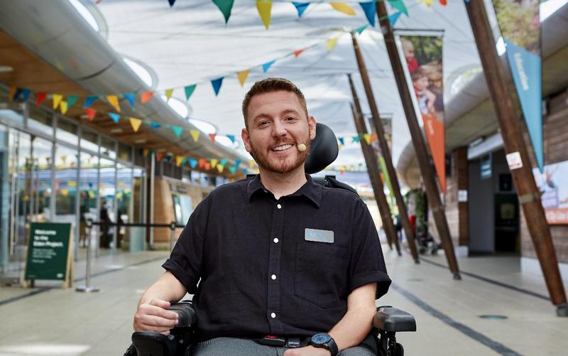 Ross sits smiling in a powered wheelchair inside a bright, modern public building decorated with colourful bunting. He wears a short sleeve black shirt and a name badge and has a small headset microphone on his cheek. Wooden beams and glass walls line the space behind him, with signs and posters visible in the background, giving the setting the feel of a busy visitor centre or venue.