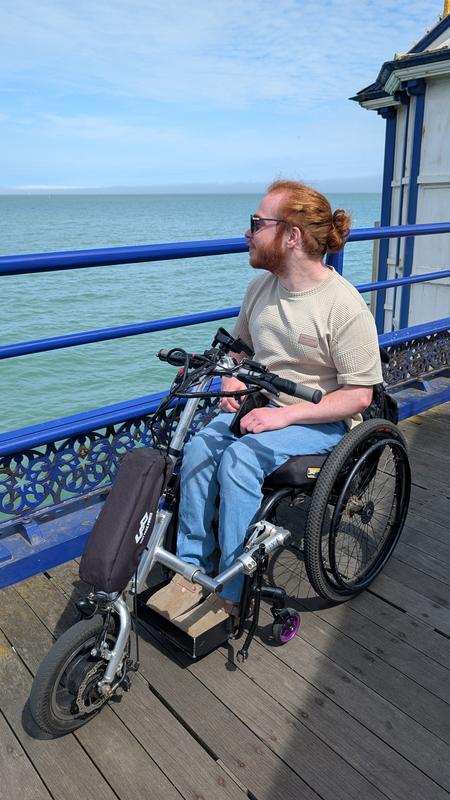 Charlie, a man with long red hair tied in a bun, sits in a wheelchair on a wooden pier, looking out over the sea. He is wearing sunglasses, a beige T shirt and light blue trousers. A handcycle attachment is fitted to the front of his wheelchair. Blue railings line the edge of the pier, with calm water and a lightly clouded blue sky in the background.