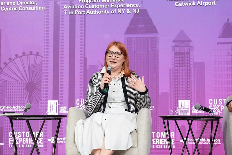 Anna Ruth sits on a stage chair speaking into a handheld microphone during a panel discussion. She wears glasses, a white dress, and a black and white jacket. A purple conference backdrop behind her shows city skyline graphics and customer experience summit branding, with small tables and microphones positioned on either side of her chair.