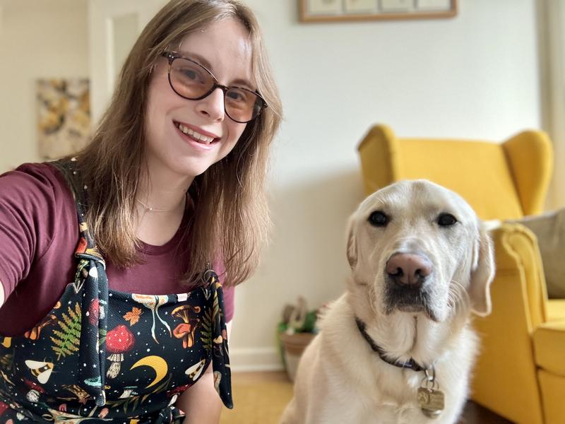 Chloe, A woman with brown hair and glasses is kneeled down next to her off duty yellow Labrador guide dog Dezzie.