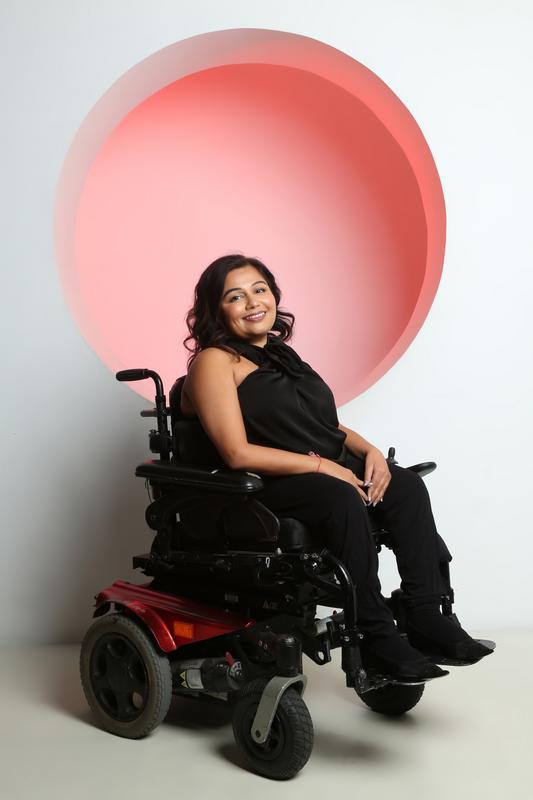 Tanvi, a woman with shoulder length dark hair, sits smiling in a red power wheelchair. She is wearing a sleeveless black outfit and black boots, angled slightly to the side with her hands resting on her lap. Behind her is a neutral studio background with a large soft red circular light.