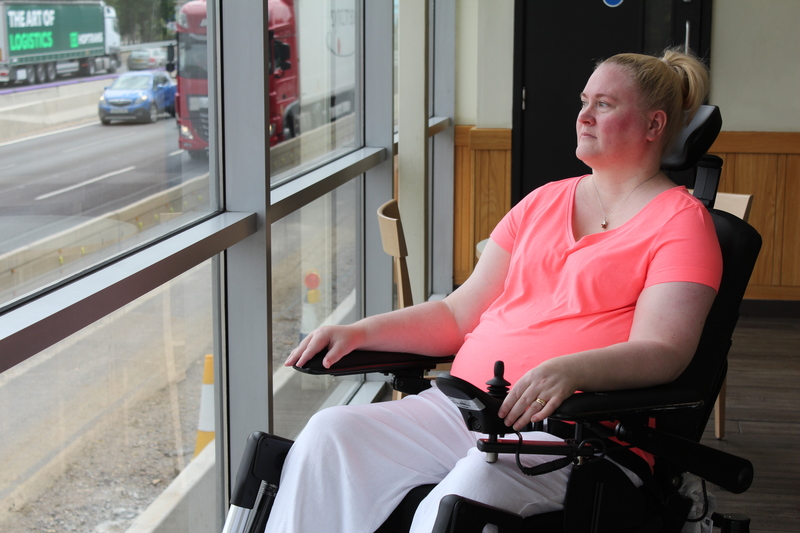 Lady with blonde hair wearing a pink t shirt, sitting in a wheelchair looking out of a window