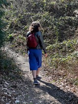 Lydia facing away from the camera and walking on a trail. She has brown hair, blue trousers and a red backpack on. 