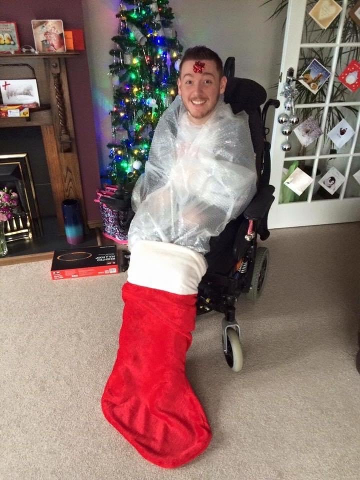 Ross sitting in his wheelchair, wrapped up with his feet in a stocking. There is a Christmas tree in the background. 