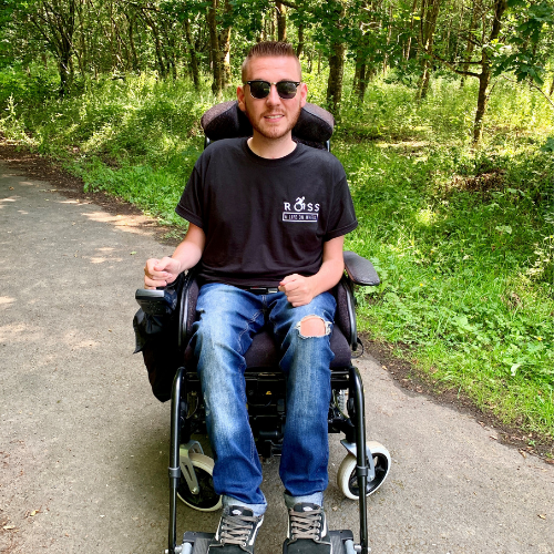 Ross is a male with brown hair and sunglasses. He is pictured in his power chair in front of greenery. He is wearing a black tshirt and blue jeans.