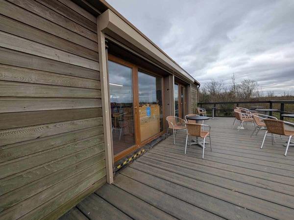 A wooden terrace with outdoor seating sits beside a building with large glass doors, overlooking surrounding vegetation under a cloudy sky