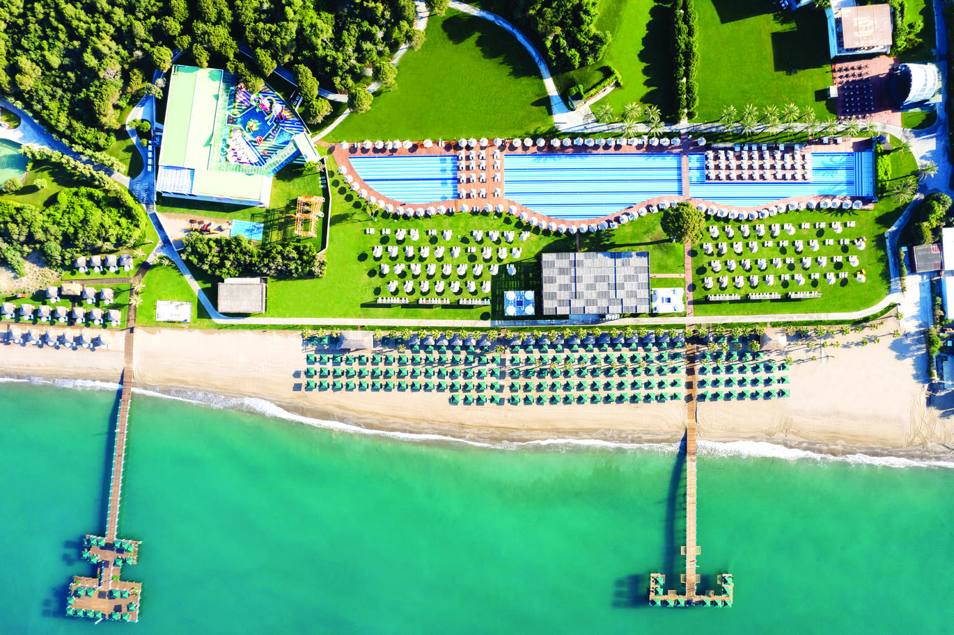 Birdseye view of the hotel complex right on the beach surrounded by lush greenery, many beds and umbrellas in the hotel and on the beach, with two piers leading out into the clear blue sea with seating areas at the front.