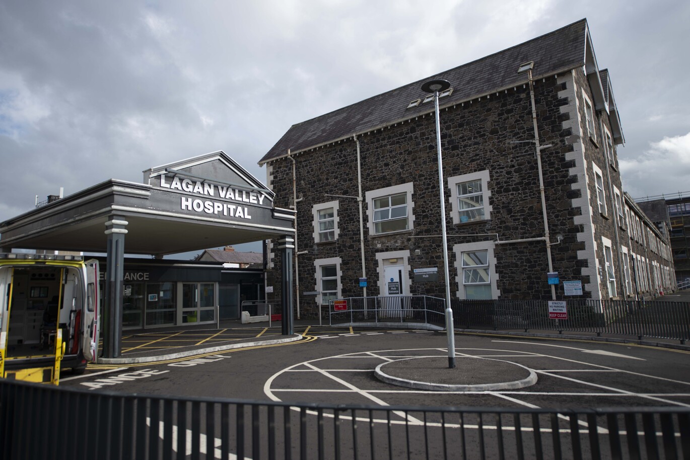 "The main entrance of Lagan Valley Hospital and COVID-19 Hub entrance. The hospital has a large canopy with 'Lagan Valley Hospital' sign at the front. The COVID-19 Hub is to the right of the main entrance. It is a three story stone building with a peaked roof. There is an ambulance to the left of the main entrance of the hospital with its back doors open and a roundabout with a lamppost in the middle at the front the hospital."