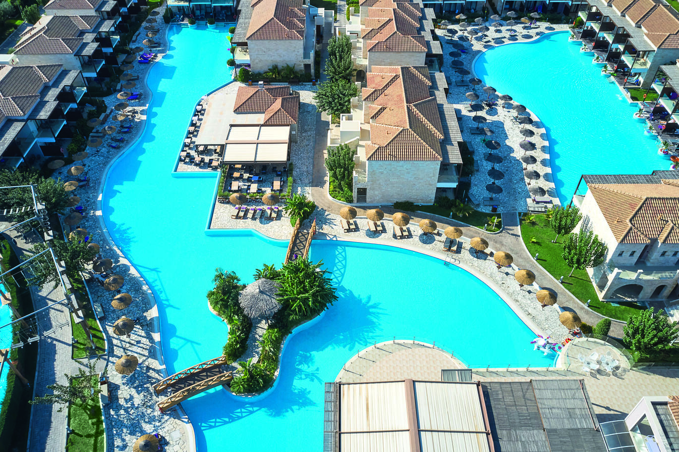 A birds eye view of the 2 swimming pools with apartment rooms surrounding and through the middle, one large with a bridge over it and one smaller on the other side of some of the apartments. Both pools are surrounded by many sun loungers and straw umbrellas with lots of greenery around.