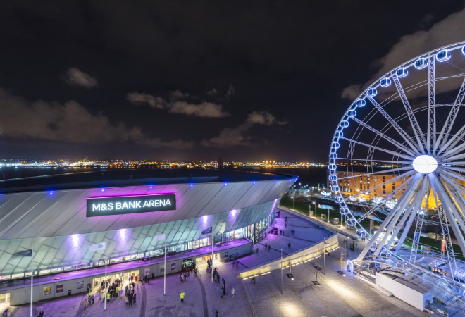 "Arial view of building lit of with blue and purple lights and wording M&S Bank Arena at the top of building. A Ferris Wheel is lit up to the right of the photo in blue lights"