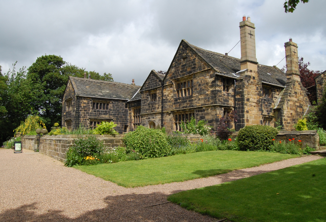 A large building made of what looks like light bricks that have become very discoloured and dark over time, there is a wall around the front which is made from the same. There are 2 large chimneys which each have a cross at the top of the bricks. There is a lovely green lawn out the front with lots of shrubbery and some poppies. At the front of the wall is a large stone gravel drive which turns into a small path between the lawn leading up to the right hand side of the lawn. There is a sign at the front, centre of the wall next to the gate.