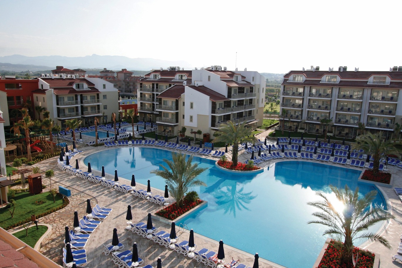 Aerial view of the pool area with many sun loungers and umbrellas around. With the hotel buildings surrounding.