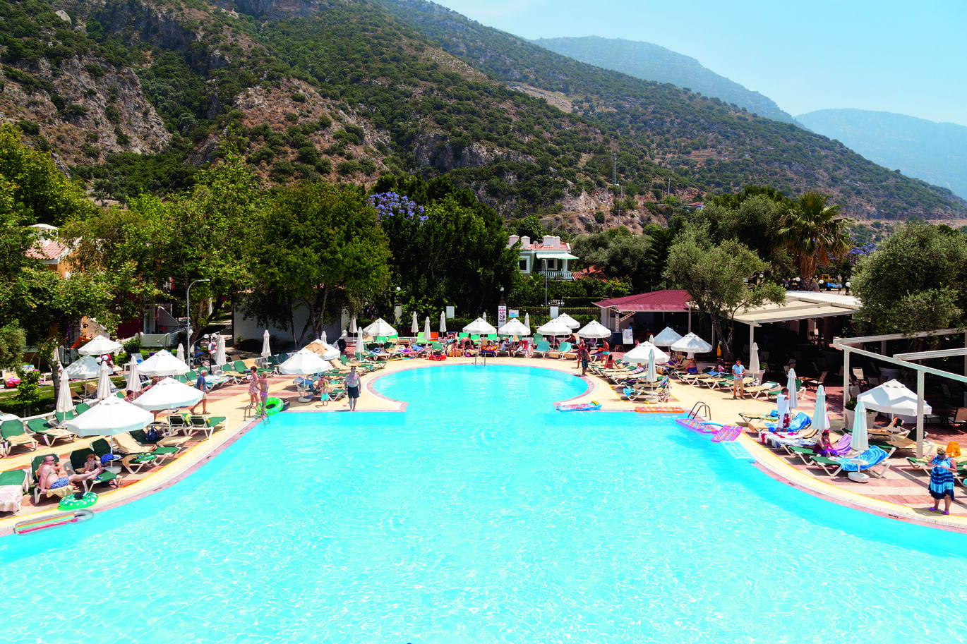 View of the pool area, surrounded by deck chairs, umbrellas and guests with trees around. With mountains behind on a clear sunny day.