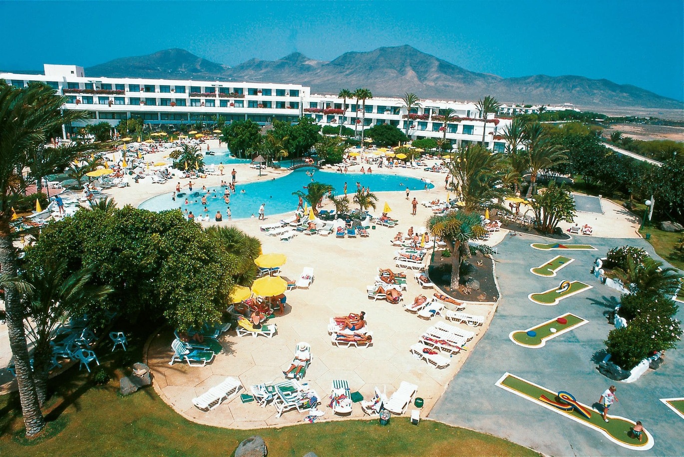 Aerial view of the pool area, surrounded by deck chairs and umbrellas with many palm trees dotted around and the hotel building behind. With mountains behind on a clear sunny day.