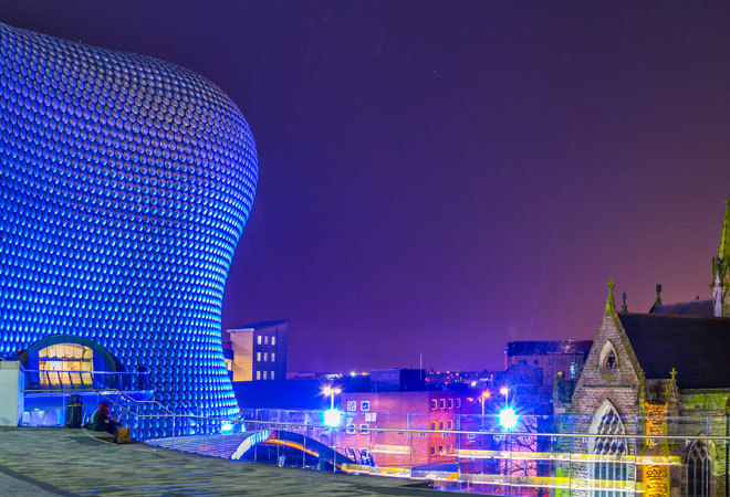 Photo of birmingham skyline at night. A modern curved building is in view, as well as a church.