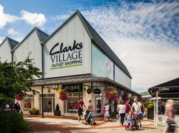 "Clarks Village outlet with shopfronts, hanging flower baskets and visitors walking along an open-air walkway."