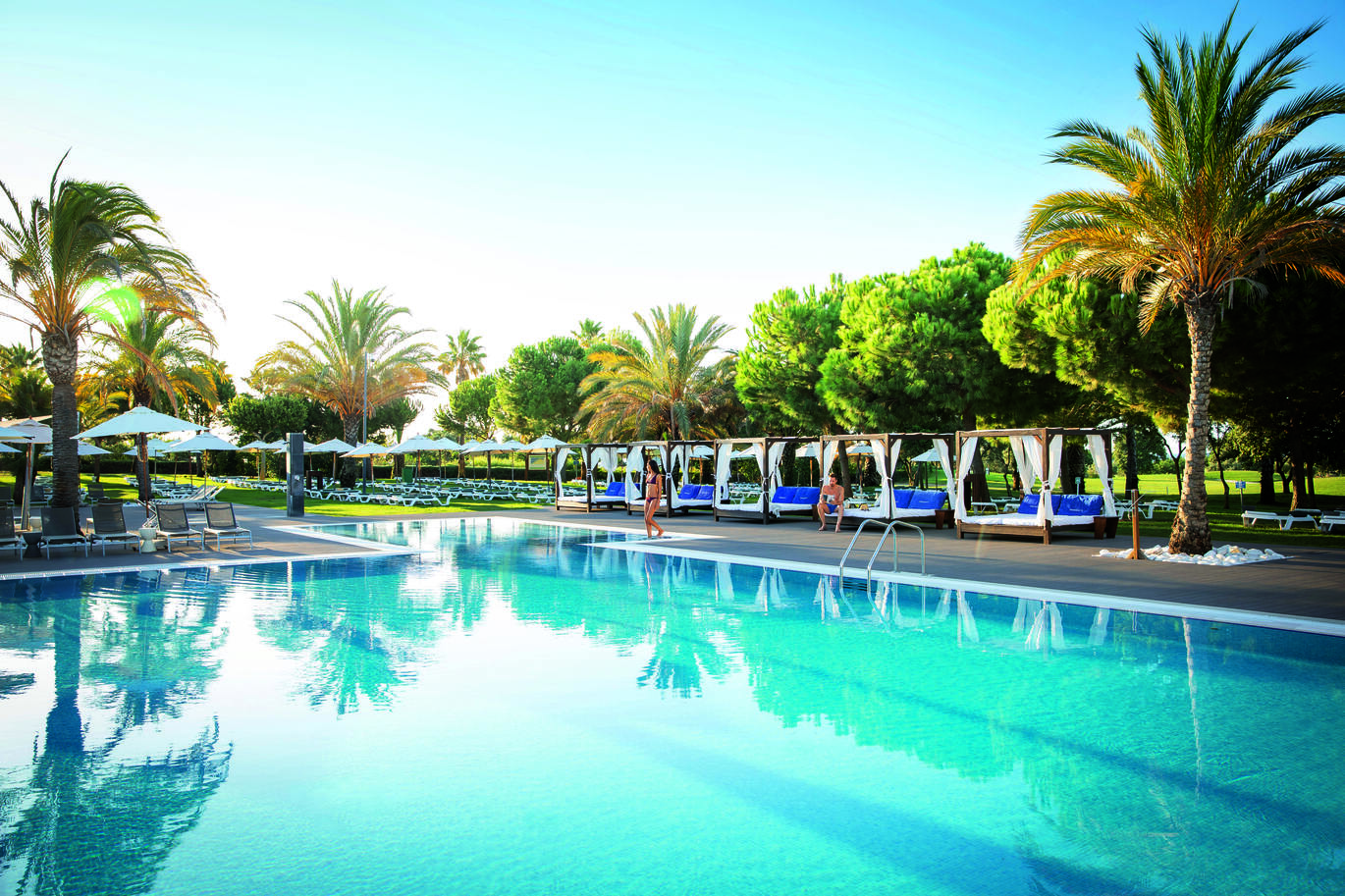 View of the pool area surrounded by sun beds and trees with a person sitting on a bed and a person walking into the pool with the blue sky above.
