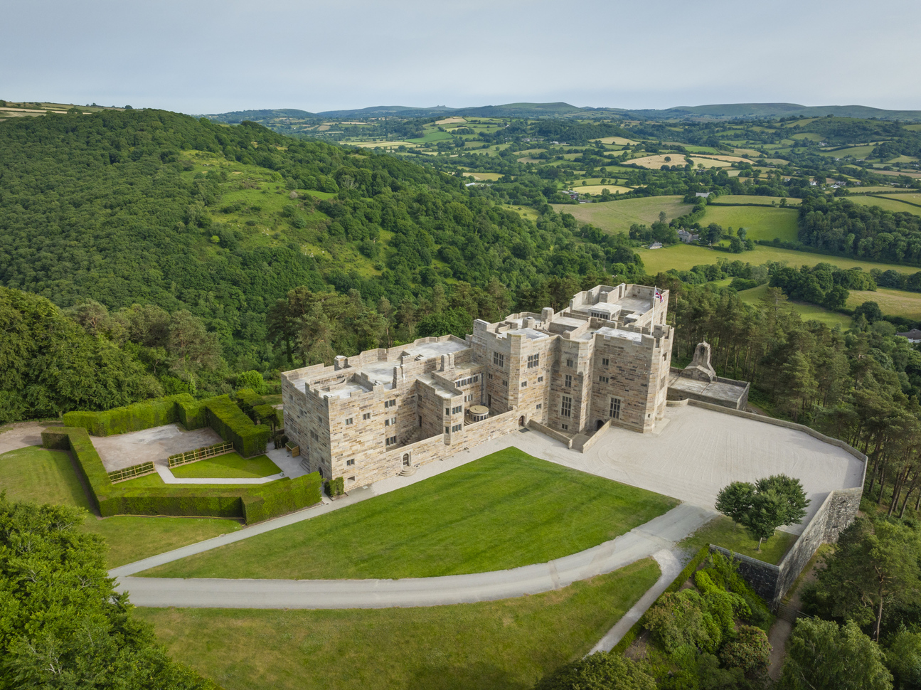 An aerial view of a castle surrounded by forests and fields. The building is very newly built with a multicoloured, light colour brick/stone, it has many windows and it looks as though you are able to walk around the roof which has steps leading onto different levels. There is a gravel driveway going in two different directions, one straight along the front of the building and one going through the lawn but both leading to the same car park. You can see a small courtyard to the right of the building and a small lawn area with a patio to the left of the building with steps down to it with a path through the lawn leading to what looks like a small car park surrounded by very neat hedges. There is an entrance to the left of the centre of the building with steps