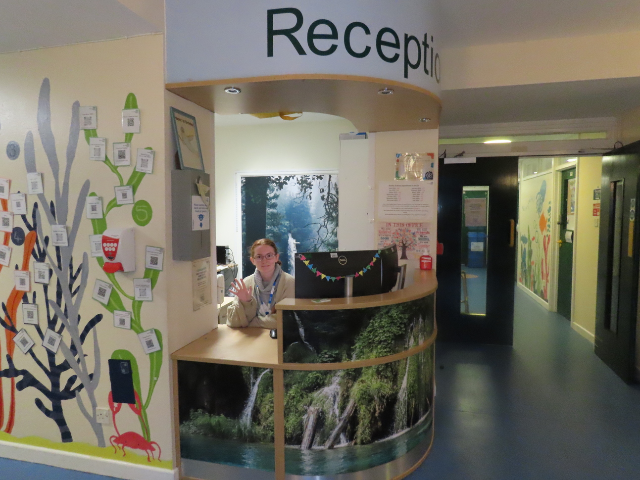 Interior of a reception area at the Plymouth Child Development Centre. A curved wooden reception desk is adorned with a nature-themed design featuring a waterfall. Behind the desk, a receptionist smiles and waves. The wall to the left is decorated with a colorful mural of tree branches, leaves, and QR codes. A hallway to the right leads to additional rooms, with walls painted in a cheerful design featuring plants and animals.