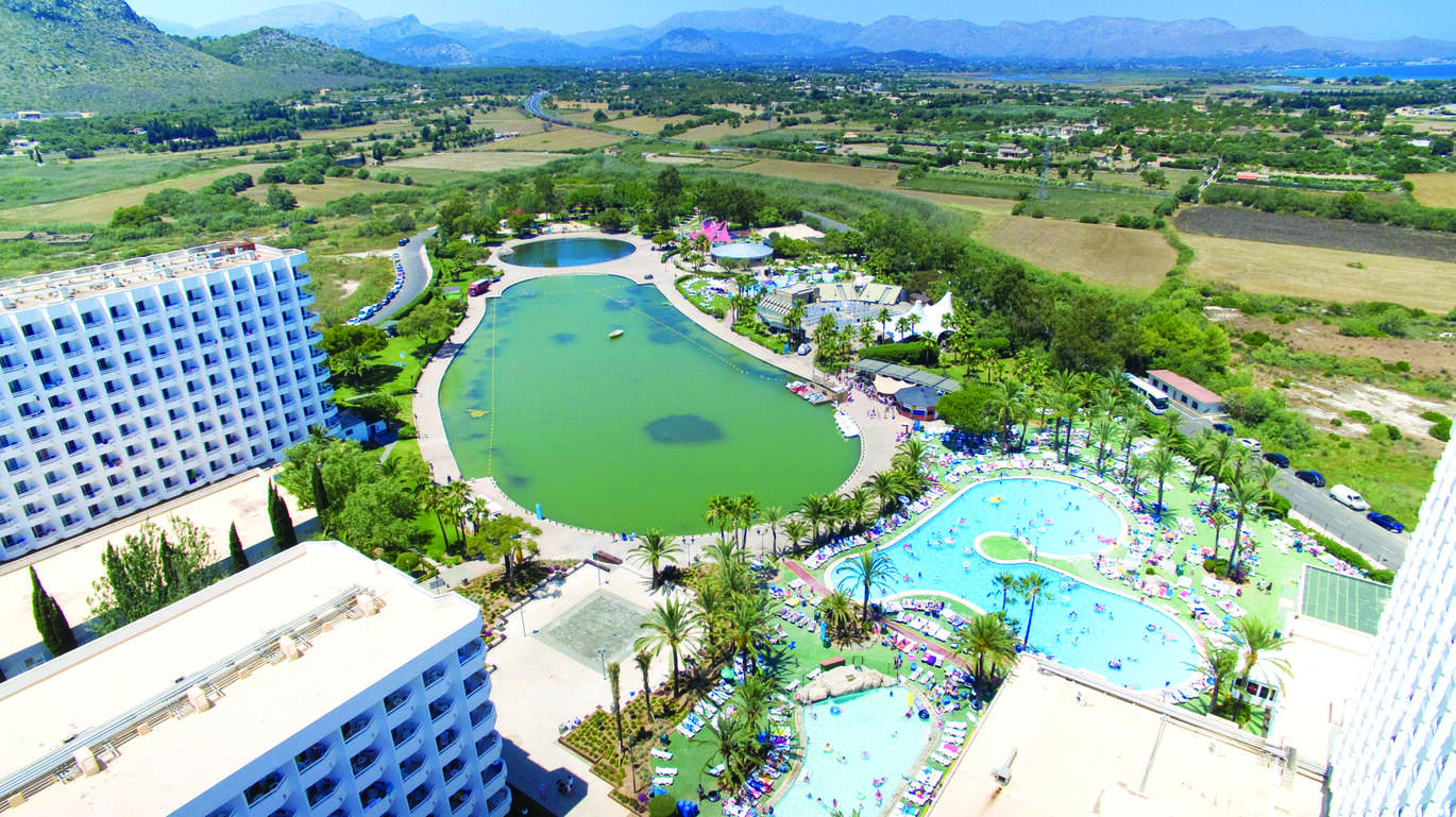 Aerial view of the hotel complex with the pools filled with people and surrounded by sun beds. The lake is behind surrounded by many fields and mountains in the background.