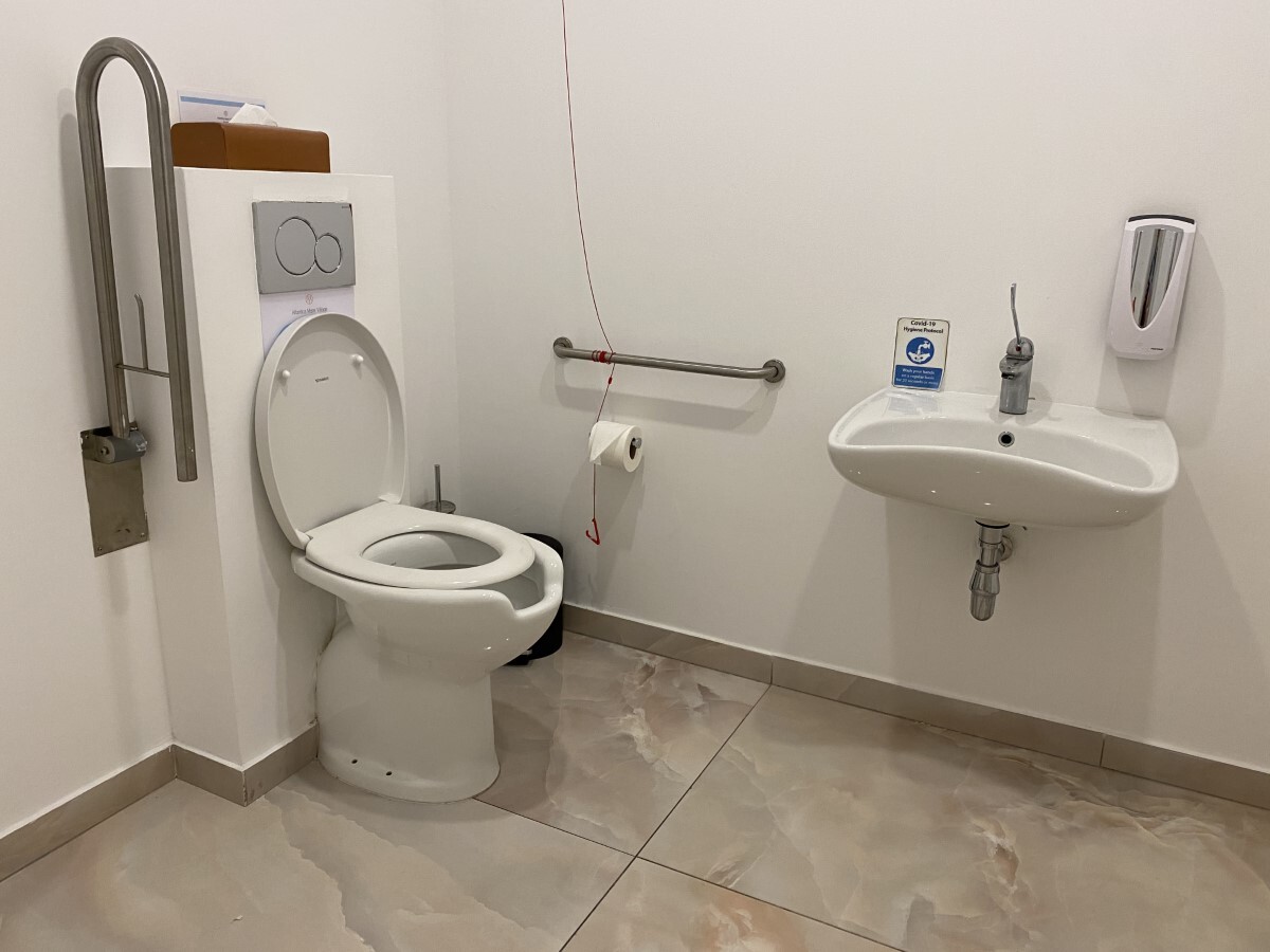 "communal toilet with adaptions for disabled people, there are grab rails and an emergency cord, toilet is left hand transfer."