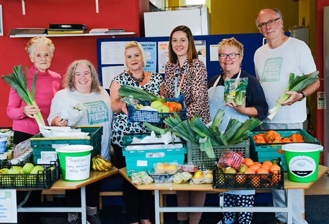 "Photo of 6 people standing behind 2 tables with lots of fruit and vegetables on.""