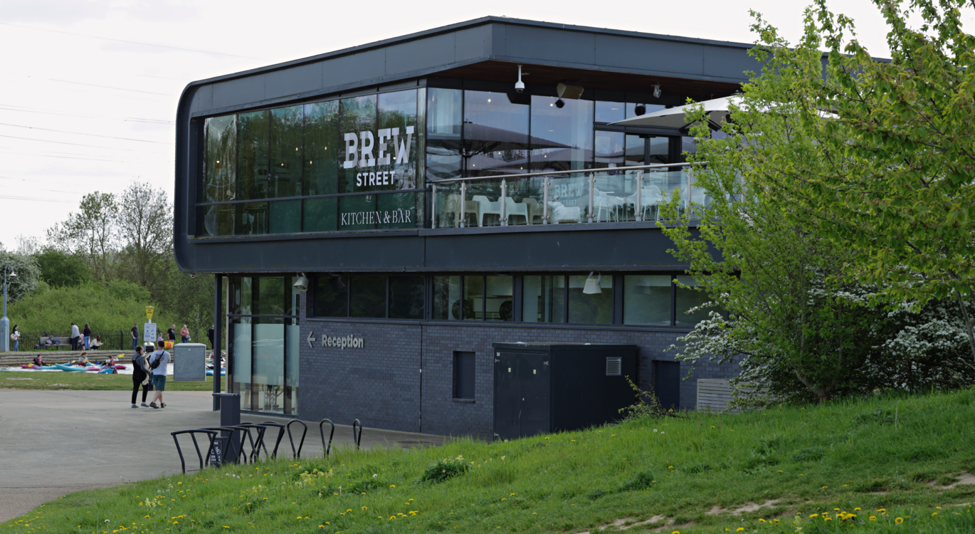 "Modern café building with glass front and outdoor terrace, beside a riverside path with people and greenery."