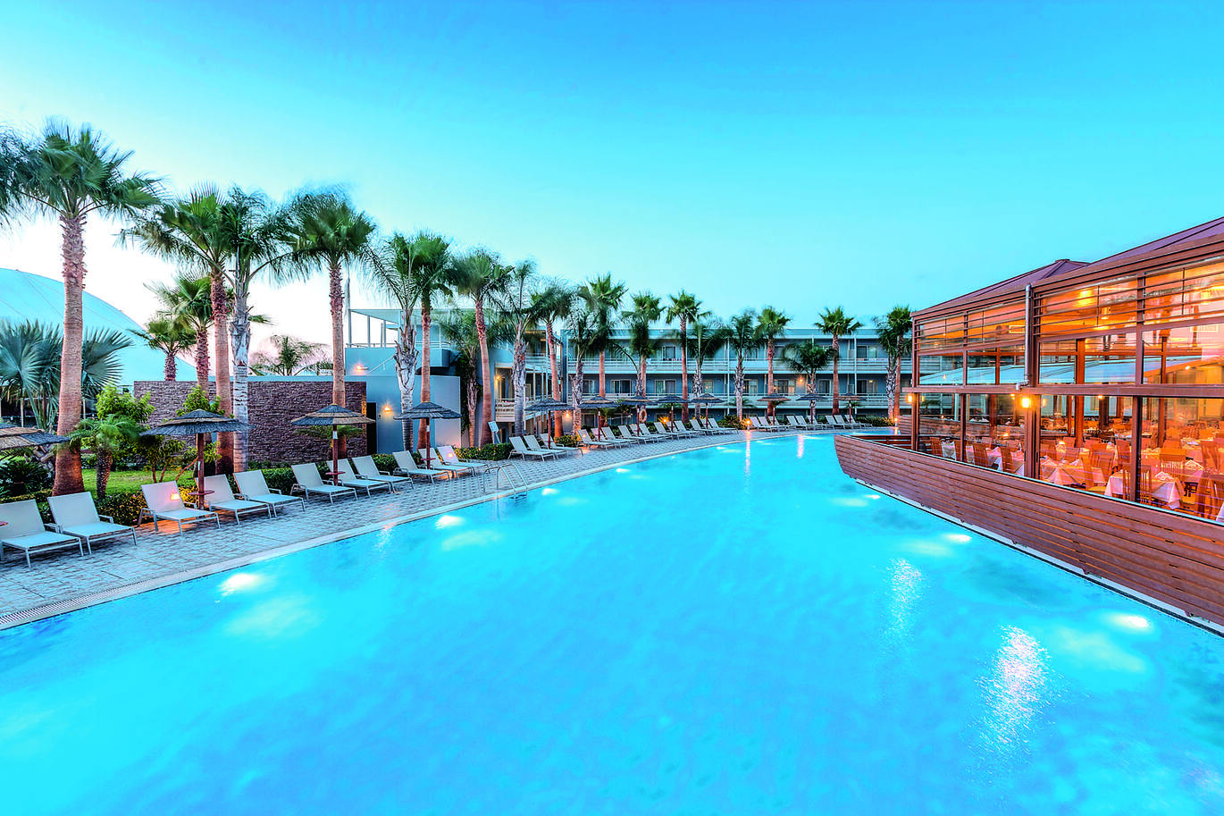 View of the pool in the sunset with sun loungers and trees to the left with clusters of hotel rooms behind, and the hotel restaurant building to the right.
