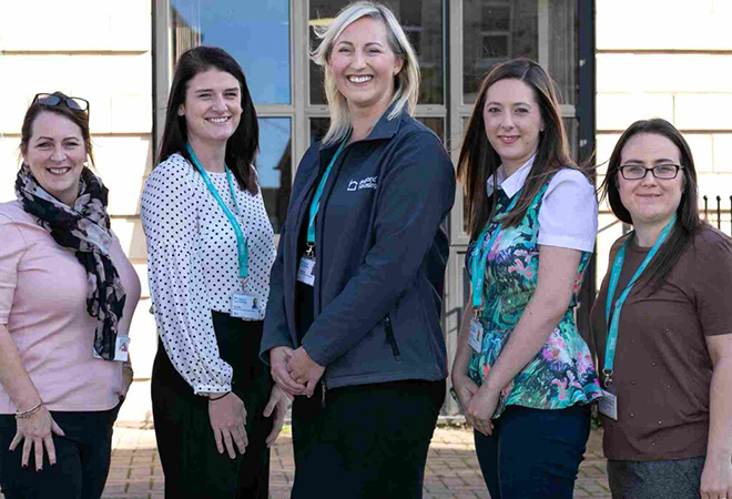 "Photo of 5 women standing for a photo outside a building. All have Beyond Housing ID tags, they are standing in height order from centre outwards."