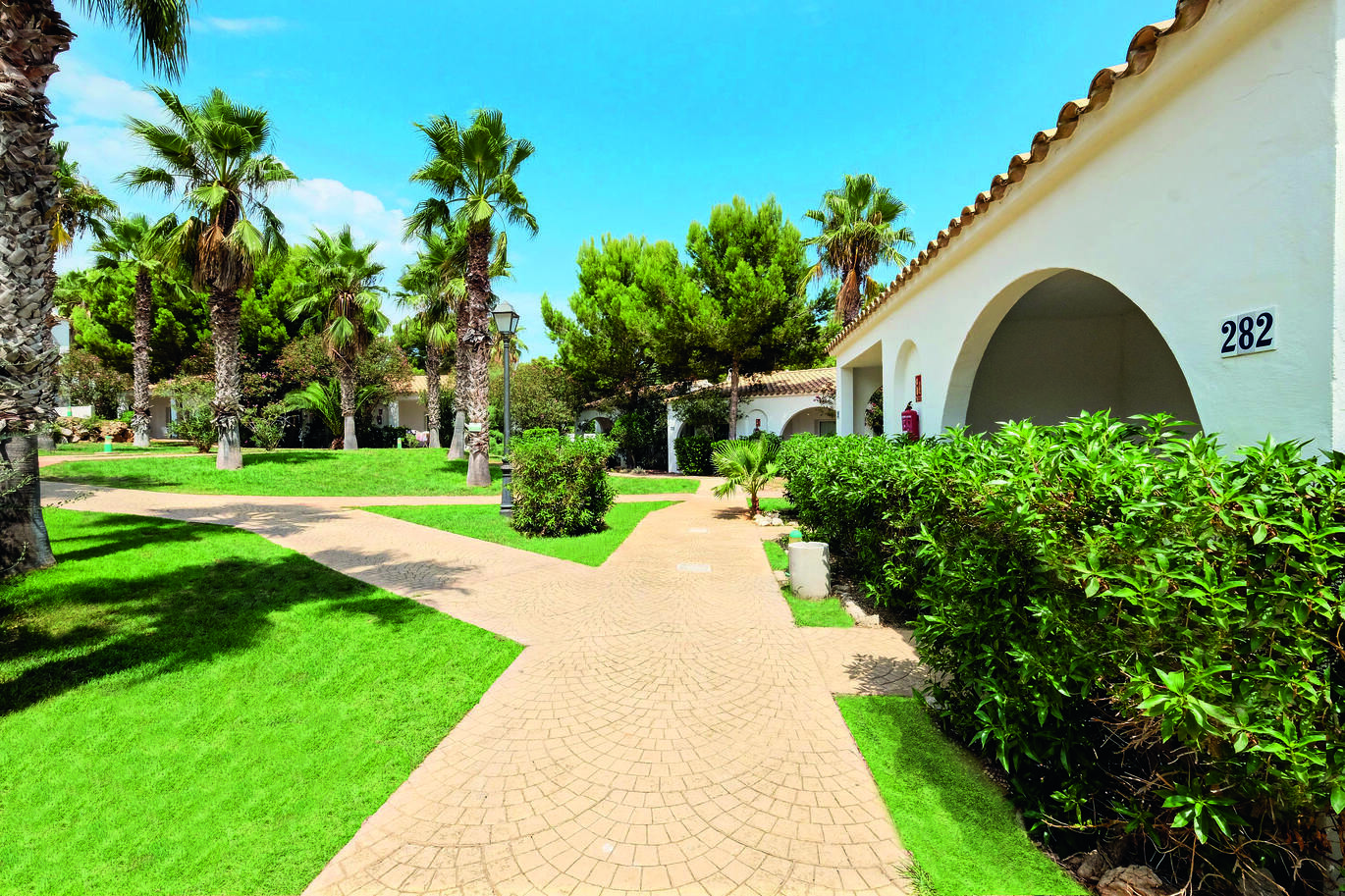 Taken from a path outside some hotel rooms with the path going off into different directions through the lush green lawn with palm trees dotted around on a lovely sunny day.
