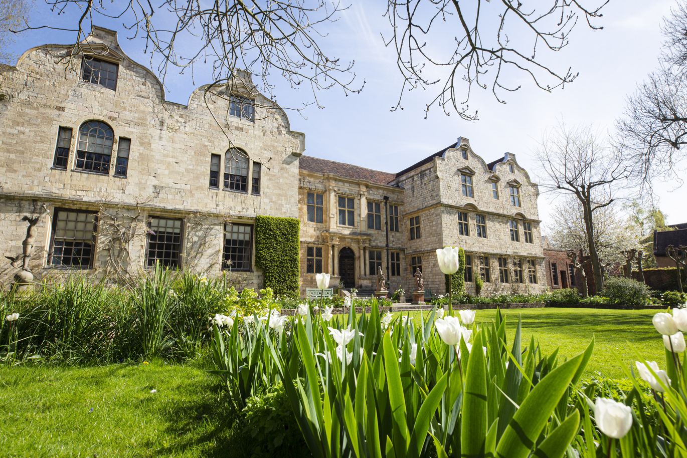 The front of the large, stone house with steps leading onto the lush green lawn surrounded by plants and flowers.