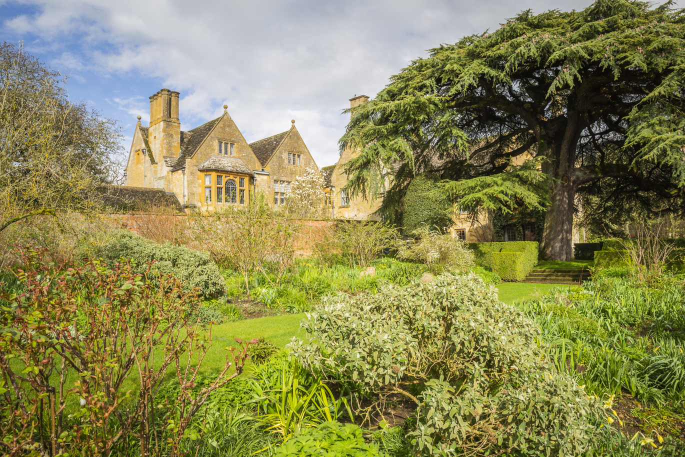 The garden in April at Hidcote full of trees, plants and greenery with a large house behind a tall brick wall.