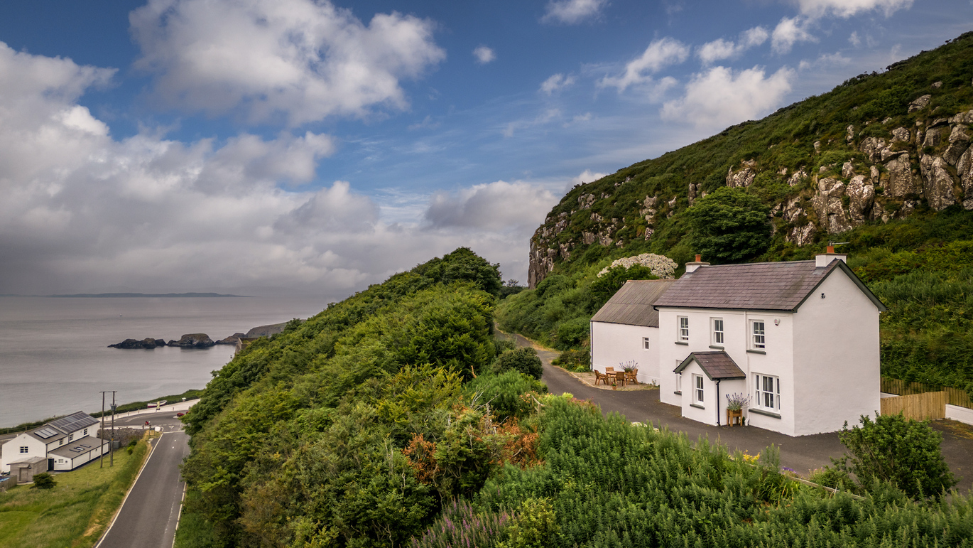 The exterior of Carrick-a-Rede Cottage, in the middle of a hill with trees below, a road below that and then the sea beyond.